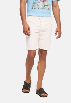 Hombre vestido con pantalones cortos blancos con cordón, camiseta azul claro con estampado tropical y sandalias negras con tiras cruzadas, de pie sobre fondo blanco.