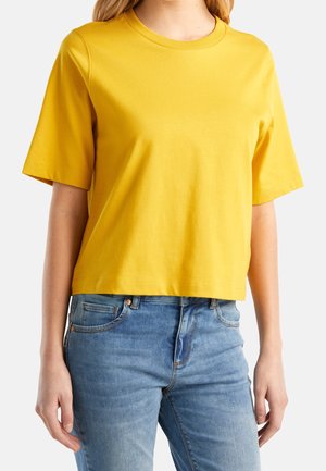 Femme portant un t-shirt jaune uni à manches courtes et un jean bleu clair sur fond blanc.