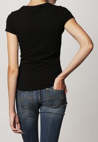 Henkilö, joka on pukeutunut istuvaan mustaan lyhythihaiseen puseroon ja sinisiin farkkuihin, pitäen yhtä kättä takapuolellaan, seisoo yksinkertaista taustaa vasten.