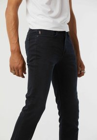Jean en denim foncé avec une coupe slim, présentant des détails de couture discrets, un design à cinq poches et une taille mi-haute. Texture lisse et marquage minimal.