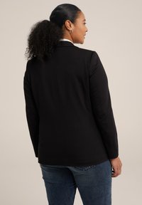 Černý blazer s přiléhavým střihem, elegantním designem a hladkou texturou. Nositý přes bílou košili, v kombinaci s modrými džíny.