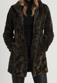 Mörkgrön fuskpälsjacka med texturerad yta, knäppning fram med tryckknappar, sidofickor och krage. Mjuk, varm och knälång design.