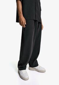 Black, loose-fitting trousers made of a smooth fabric, paired with white sneakers featuring grey accents. Includes a relaxed silhouette and minimalistic design.