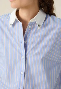 Chemise rayée bleu clair avec col blanc, présentant un matériel décoratif aux pointes du col. Tissu doux, devant à boutons et rayures verticales.