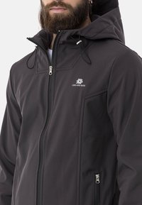 Schwarze Softshelljacke mit einem Reißverschluss vorne, verstellbarer Kapuze und seitlichen Taschen. Mit einem Logo auf der Brust und Kontrastnähten.