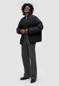 Czarna kurtka puchowa z zamkiem błyskawicznym, szerokim fasonem, gładką teksturą i naszywką z logo. Połączona z szarymi spodniami o szerokich nogawkach i czarnymi butami.