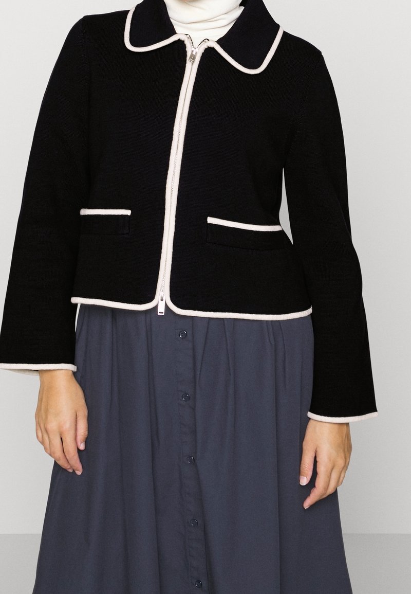 Veste noire zippée avec bordure blanche, portée sur un col roulé blanc et une jupe gris foncé à boutons.