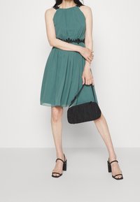 Smaragdgrön ärmlös klänning med samlad midja och svart blommig broderi. Svart texturerad handväska och svarta sandaletter med blockklack.