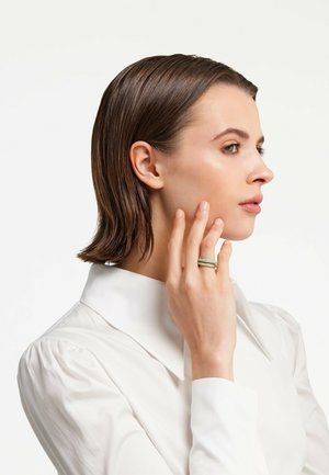 Stilvoller Ring mit einem rechteckigen grünen Stein und goldfarbenen Akzenten, getragen am Zeigefinger einer Person in einem weißen Hemd mit Kragen.