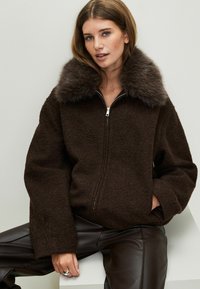 Chaqueta de forro marrón con un cuello de piel, cierre de cremallera, hombros caídos y mangas anchas. Combinada con pantalones de cuero marrón oscuro.