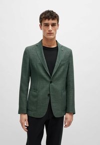 Blazer verde con texture, con revers a punta, due tasche frontali e chiusura con un solo bottone, indossato sopra una camicia nera.