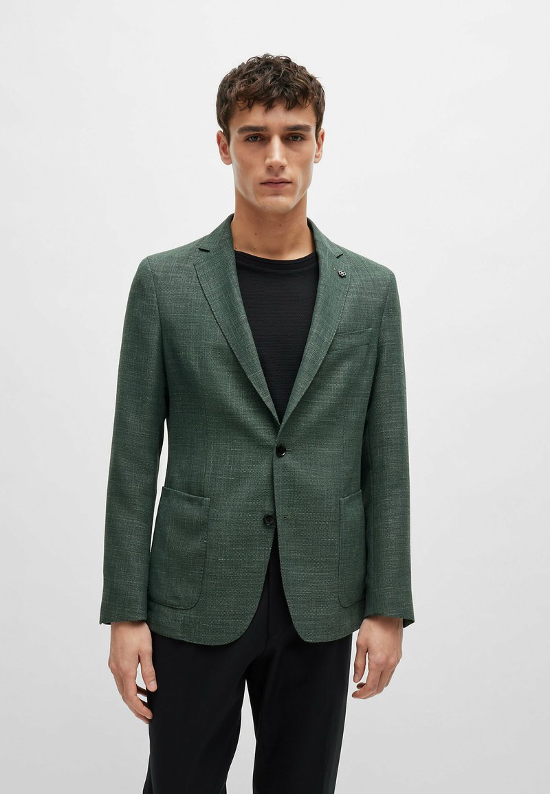 Blazer verde con texture, con revers a punta, due tasche frontali e chiusura con un solo bottone, indossato sopra una camicia nera.