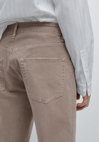 Jasnobrązowe dżinsy denimowe mają dopasowany krój i dwie tylne kieszenie. Osoba nosi białą koszulę z długim rękawem w paski.