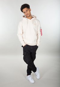 Hoodie beige avec poche avant et logo, associé à un pantalon cargo noir avec des poches latérales, et des baskets blanches avec une semelle texturée.