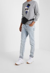Uomo che indossa jeans azzurri, sneakers bianche, felpa grigia con grafica di una persona in cappello e occhiali da sole, e una borsa a vita nera.