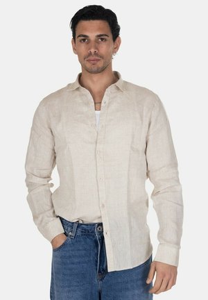 Uomo con capelli scuri che indossa una camicia beige con bottoni sopra una maglietta bianca e jeans blu, in piedi con una mano in tasca.