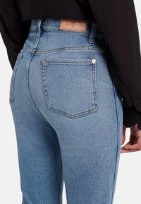 Jeans in denim blu chiaro con vita alta, dotati di un'etichetta beige sulla cintura posteriore e due tasche posteriori, texture liscia con dettagli minimi.
