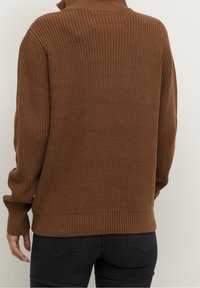 Pull à col roulé marron tricoté porté par une personne vue de dos, associé à un jean noir ajusté sur un fond uni.