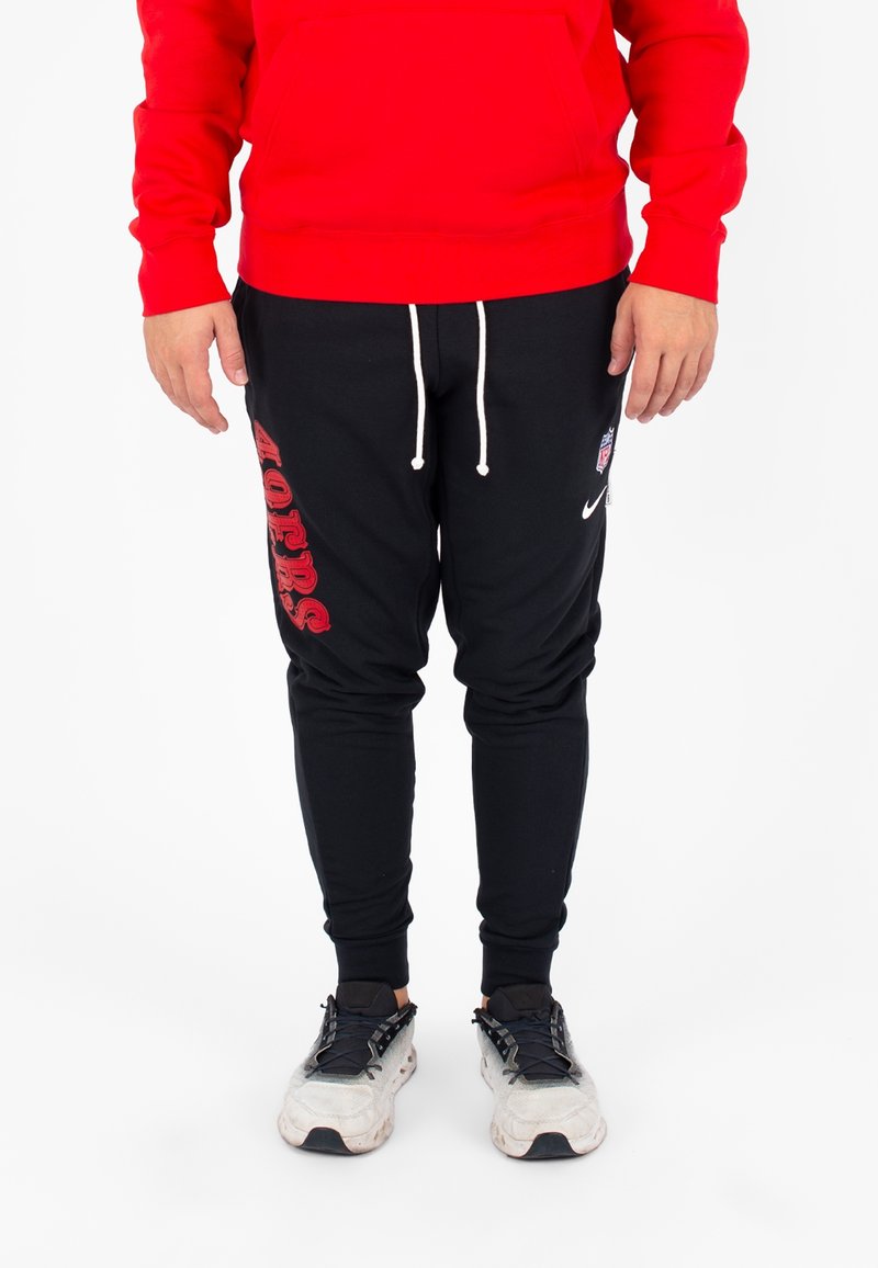 Pantaloni sportivi neri con stampa rossa "49ers" sulla gamba sinistra, cordino bianco e caviglie elastiche, abbinati a una felpa rossa e scarpe da ginnastica grigie.