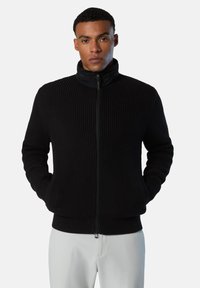 Schwarzer, gerippter Zip-Up-Pullover mit hohem Kragen, strukturiertem Stoff und taillierter Form, getragen mit hellen Hosen.