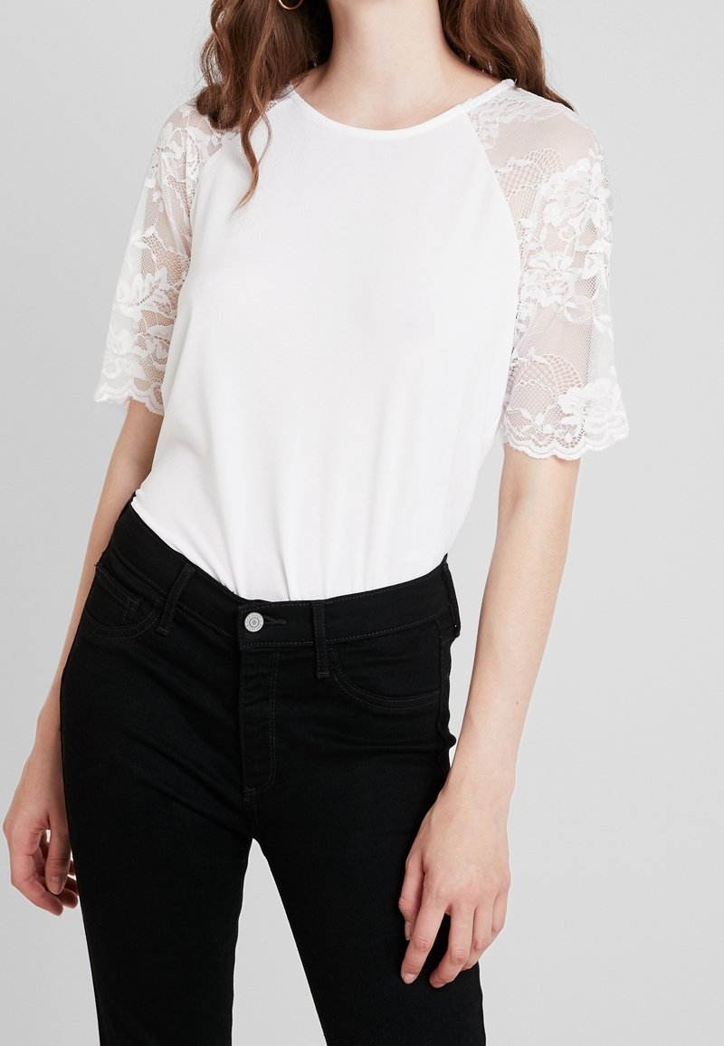 Vit kortärmad topp med spetsdetaljer på ärmarna, kombinerad med svarta jeans. Slätt tyg med rund halsringning och figurnära passform.