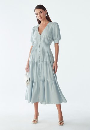 Femme portant une robe midi bleu clair à boutons sur le devant avec des manches ballon, tenant un sac à main blanc et portant des sandales blanches à talons.