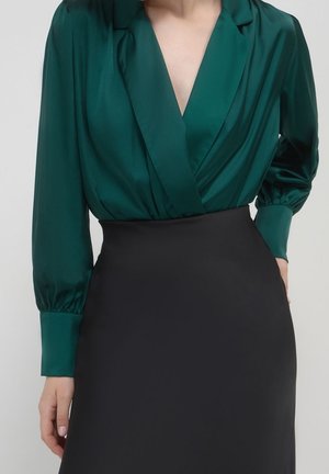 Kvinde iført en dybgrøn satinbluse med lange puffede ærmer og en sort nederdel med høj talje, vist fra nakke til hofter.