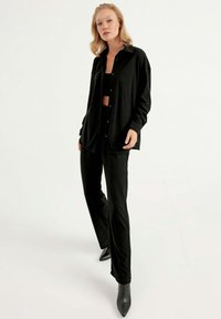 Schwarzes Hemd mit Knopfleiste aus strukturiertem Stoff, kombiniert mit eng anliegenden schwarzen Hosen. Beide Teile haben einen lässigen Schnitt und ein minimalistisches Design.