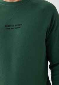 Sweatshirt vert en tissu doux, avec un col ras du cou, des manches raglan et un texte brodé "CRAFTED GOODS - PIER ONE DENIM".