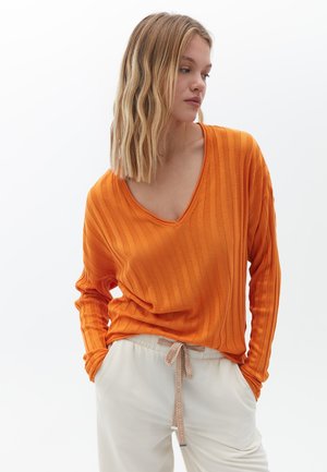 Oranje geribbelde lange mouwen top met een brede V-hals. Gecombineerd met crèmekleurige broek met een geprinte trekkoord. Gladde textuur.