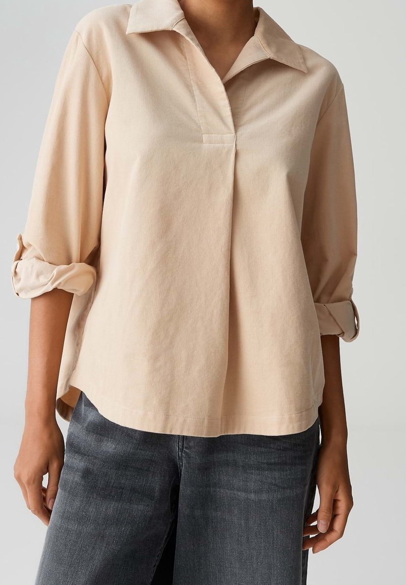 Chemise beige à manches longues avec poignets retroussés portée avec un jean gris foncé sur une personne debout, cadrée au-dessus des épaules et des genoux.