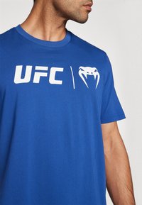 Royal blue bomulds T-shirt med hvidt trykt "UFC"-tekst og kobra logo. Korte ærmer og rund halsudskæring. Glat tekstur, letvægtsdesign.