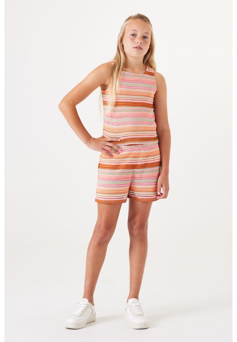 Jong meisje staat zelfverzekerd met één hand op de heup, gekleed in een gestreepte mouwloze top en bijpassende shorts met witte sneakers.
