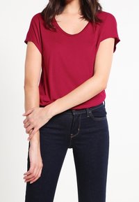 Kvindelig iført en kortærmet rød T-shirt og mørkeblå jeans, med armene korslagt foran et hvidt baggrund.