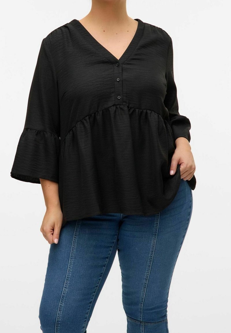 Femme portant un chemisier noir ample avec trois boutons et des manches évasées, associé à un jean skinny bleu.