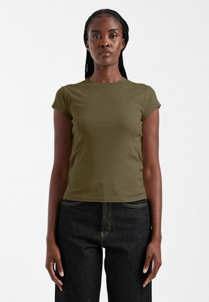 Femme aux cheveux tressés portant une chemise ajustée à manches courtes de couleur vert olive et un jean noir taille haute, debout devant un fond blanc.