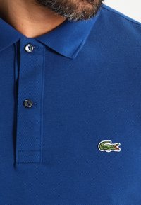 Blue polo shirt made of textured fabric, featuring a two-button placket and a small green crocodile logo embroidered on the chest.