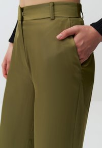 Oliwkowe, wysokie spodnie o gładkiej fakturze, z bocznymi kieszeniami i gładkim przodem. Widoczne subtelne detale szycia.
