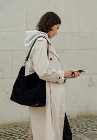 Beige trench coat layered over a gray hooded sweatshirt, holding pamphlets. Black suede tote bag with a curved shape and shoulder strap.