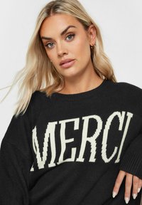 Svart stickad tröja med stort "MERCI"-tryck i krämfärgat. Rund halsringning och sänkt axel med en strukturerad yta.