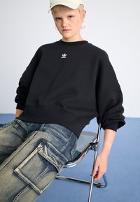Juoda didelio dydžio sportinė palaidinė su guma aptrauktomis rankovėmis, su mažu baltu Adidas logotipu. Derinama su šviesiai mėlynomis kargo džinsomis, turinčiomis dideles kišenes.