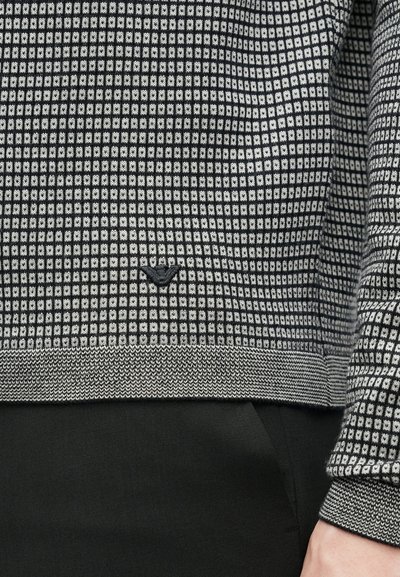 Kötött pulóver fekete-fehér kockás mintával, bordázott mandzsettákkal és szegéllyel, valamint egy kis logóval az eleje alján.