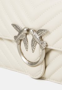 Borsa a tracolla in pelle crema trapuntata con clasp in metallo che presenta due uccelli che si baciano e un anello circolare. Motivo a zigzag cucito sulla superficie.
