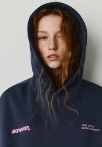 Giovane donna con lunghi capelli rossi indossa una felpa con cappuccio blu navy, cappuccio alzato, con il testo rosa "STWD*" e "STAY UNITED GLOBAL COMMUNITY" sul davanti.