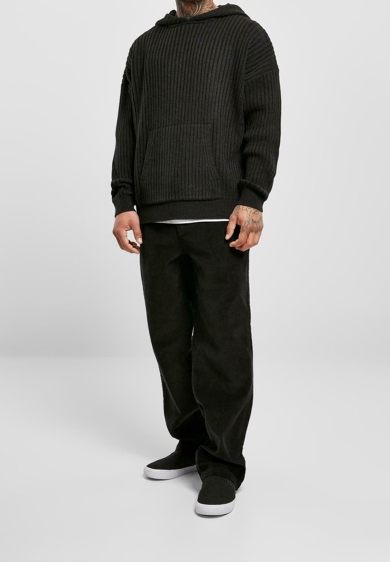 Homme portant un sweat à capuche noir côtelé, pantalon noir ample, chaussures slip-on noires avec semelles blanches, debout devant un fond clair uni.