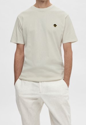 Beżowa koszulka z okrągłym dekoltem z miękkiej bawełny, z małym czarno-żółtym logo na piersi, zestawiona z jasnymi lnianymi spodniami.