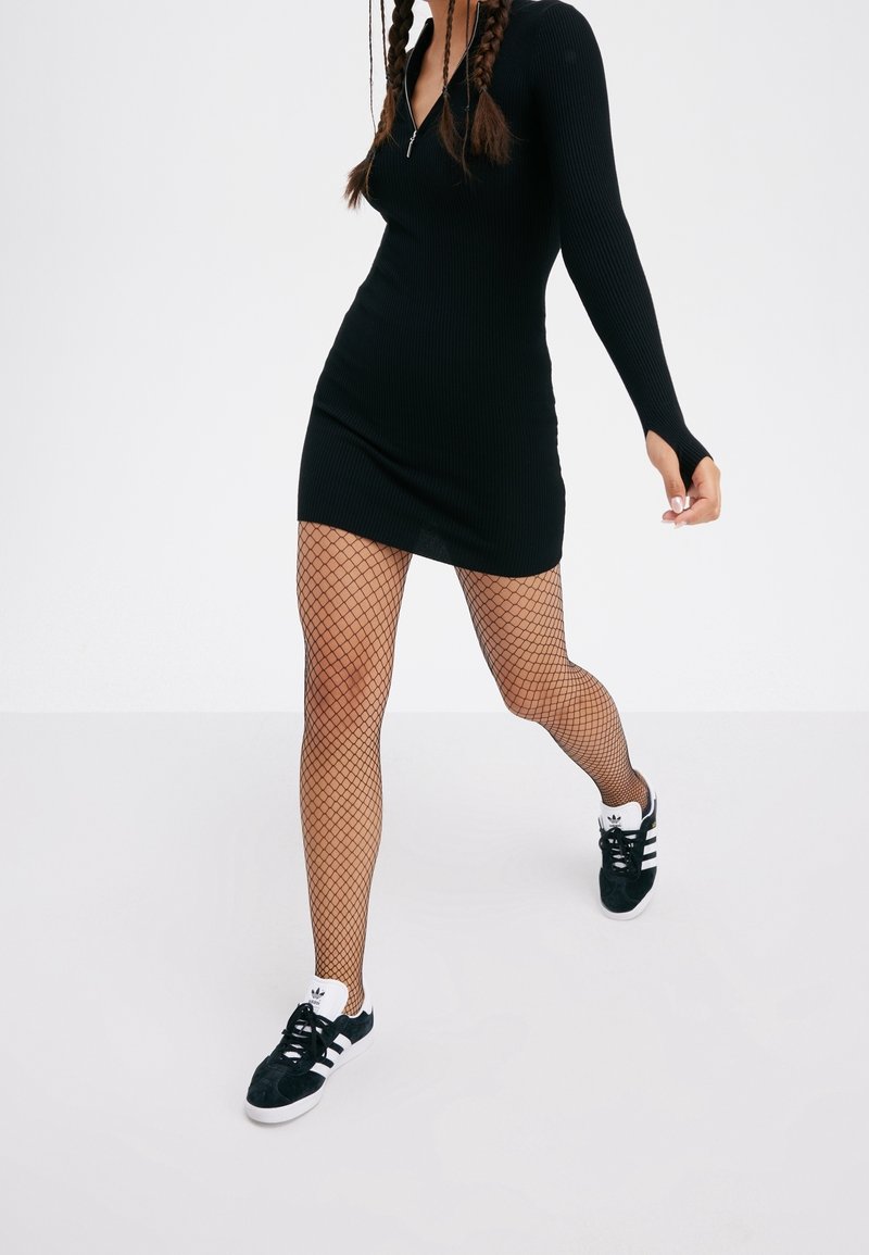 Mini abito nero a costine con maniche lunghe, caratterizzato da una zip al collo. Abbinato a collant a rete e sneakers in suede nere.