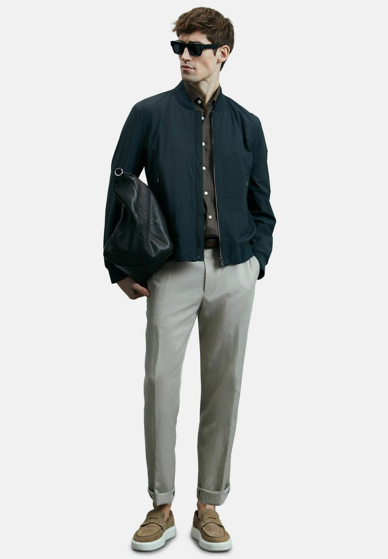 Mann in schwarzer Bomberjacke, braunem Hemd, hellgrauen Hosen, hellbraunen Slippern, Sonnenbrille, hält eine schwarze Ledertasche und steht vor einem weißen Hintergrund.