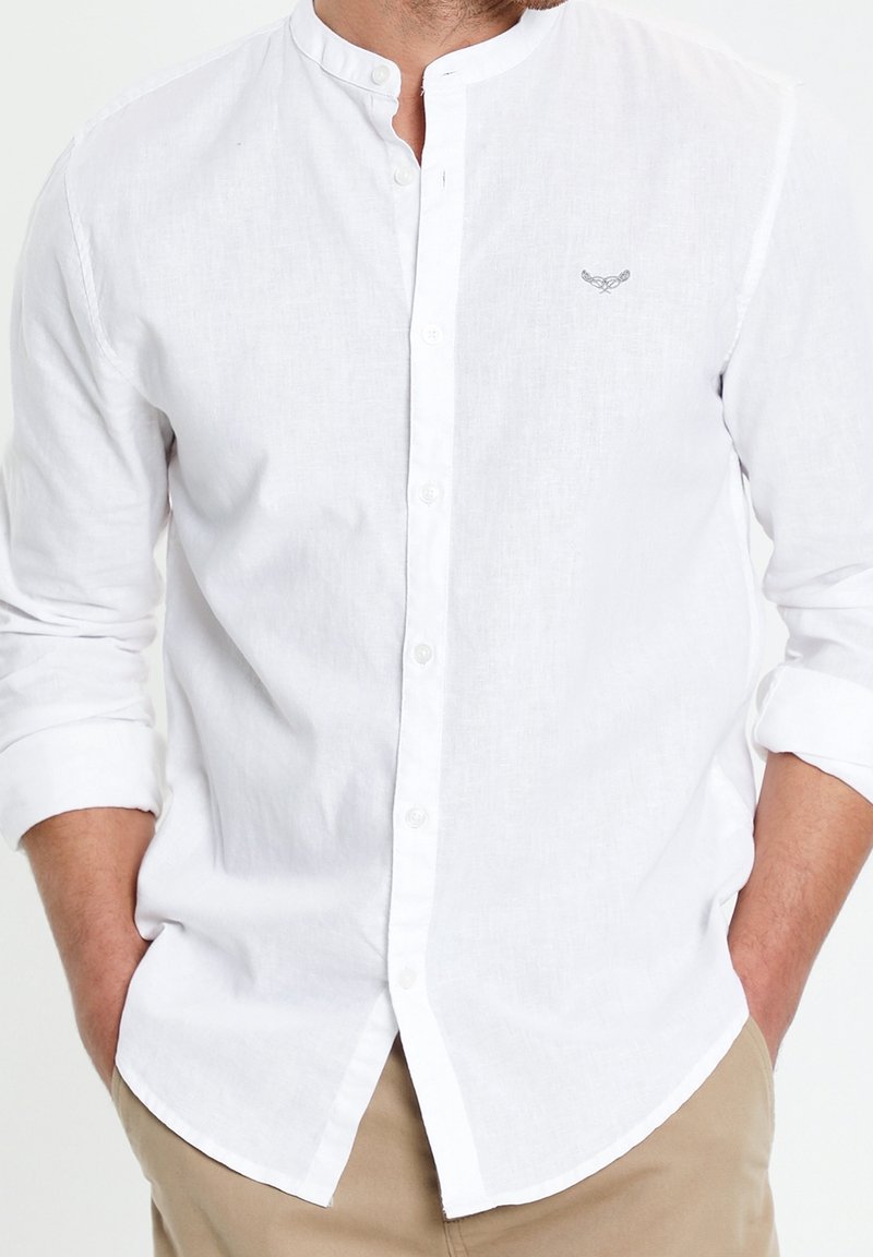 Chemise en lin blanc avec col mandarin, manches longues retroussées aux poignets, devant boutonné et un subtil détail brodé sur la poitrine.