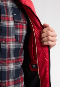 Rote isolierte Jacke mit glattem Äußeren und beige Absetzungen, teilweise offen, um ein navy- und rotkariertes Hemd mit braunen Knöpfen zu zeigen.